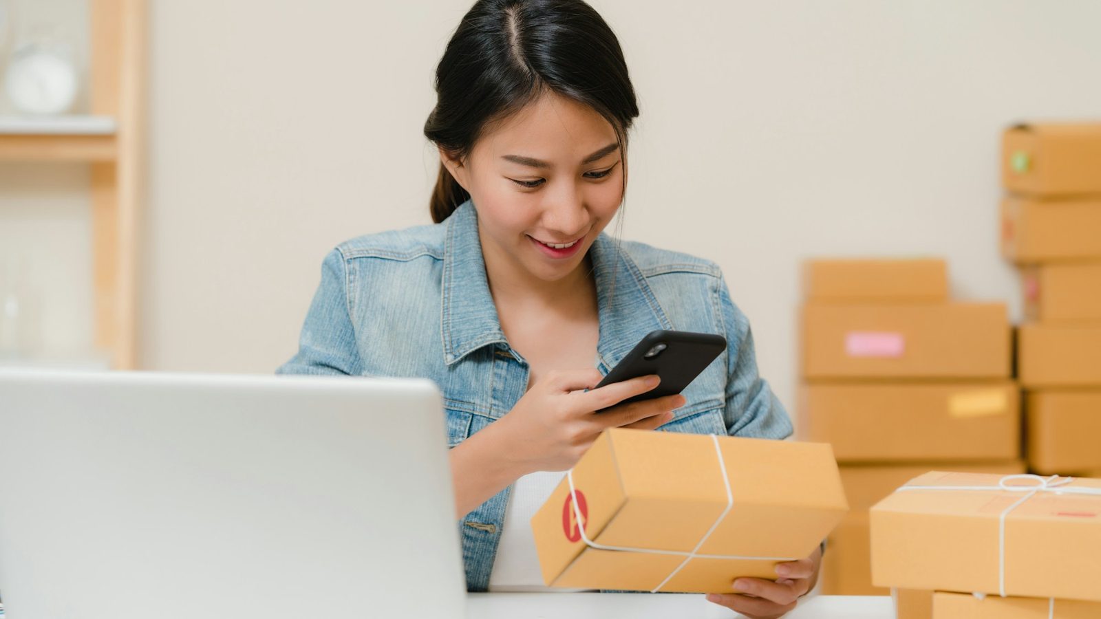 A woman checking a parcel with her phone, boxes behind her