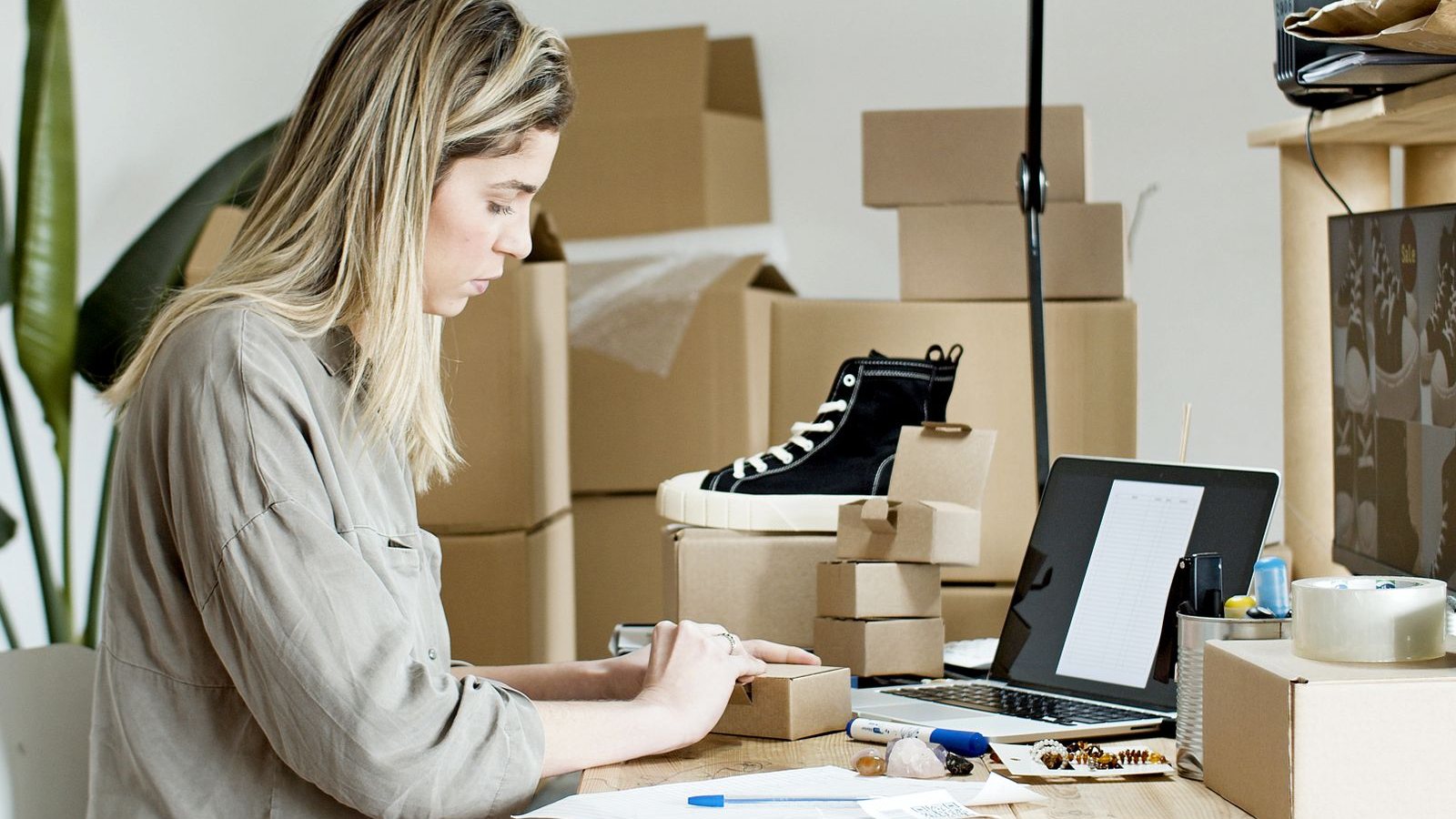 An online shop owner packing an order at her desk, with a laptop and boxes ready to ship